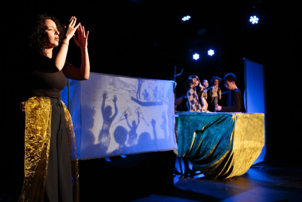 British Sign Language Interpreter Sumayya Si-Tayeb signing in front of a puppet screen showing disabled people dancing at a rave. In the background, puppeteers Gun Suen, Sara Guedesh and Raven Kaliana operate table-top puppets.