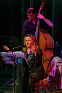 Reem Kelani sitting on stage, singing, with her arms raised. A man is playing doubel bass behind her.