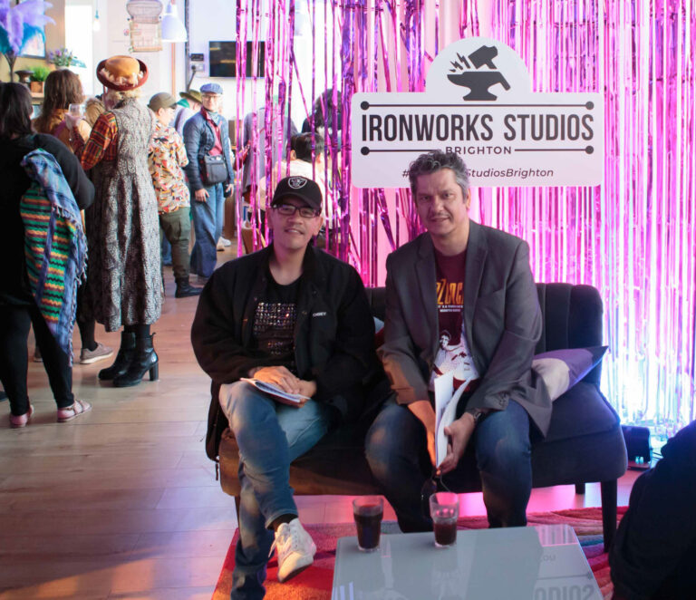 Access Consultant Troi Lee of Deaf Rave and Dramaturg Hassan Mahamdallie, sitting in front of a pink curtain in the Ironworks Studios foyer