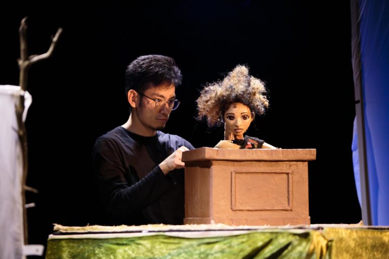 Puppeteer Gun Suen with puppet Peat, a young man with a large Afro sitting in a witness stand